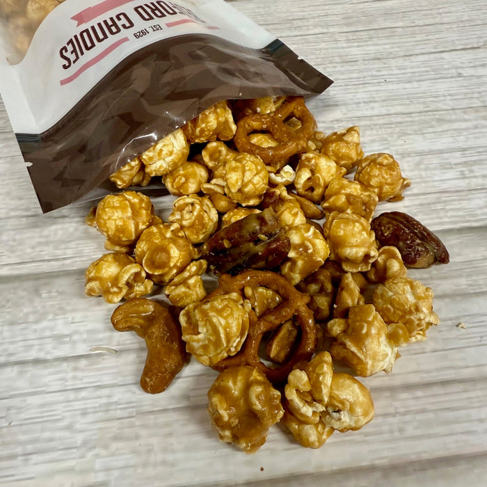 Caramel popcorn with pretzels and nuts spilling out of a bag on a wooden surface.